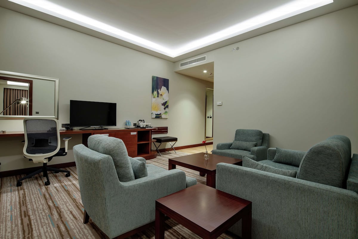A modern hotel room with a flat-screen TV, grey upholstered sofas, a wooden coffee table, a desk with a computer, and a large mirror.