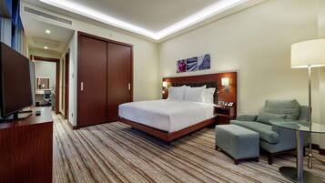 A hotel room with a bed, a desk with a TV, a chair, a glass table, and a wardrobe.