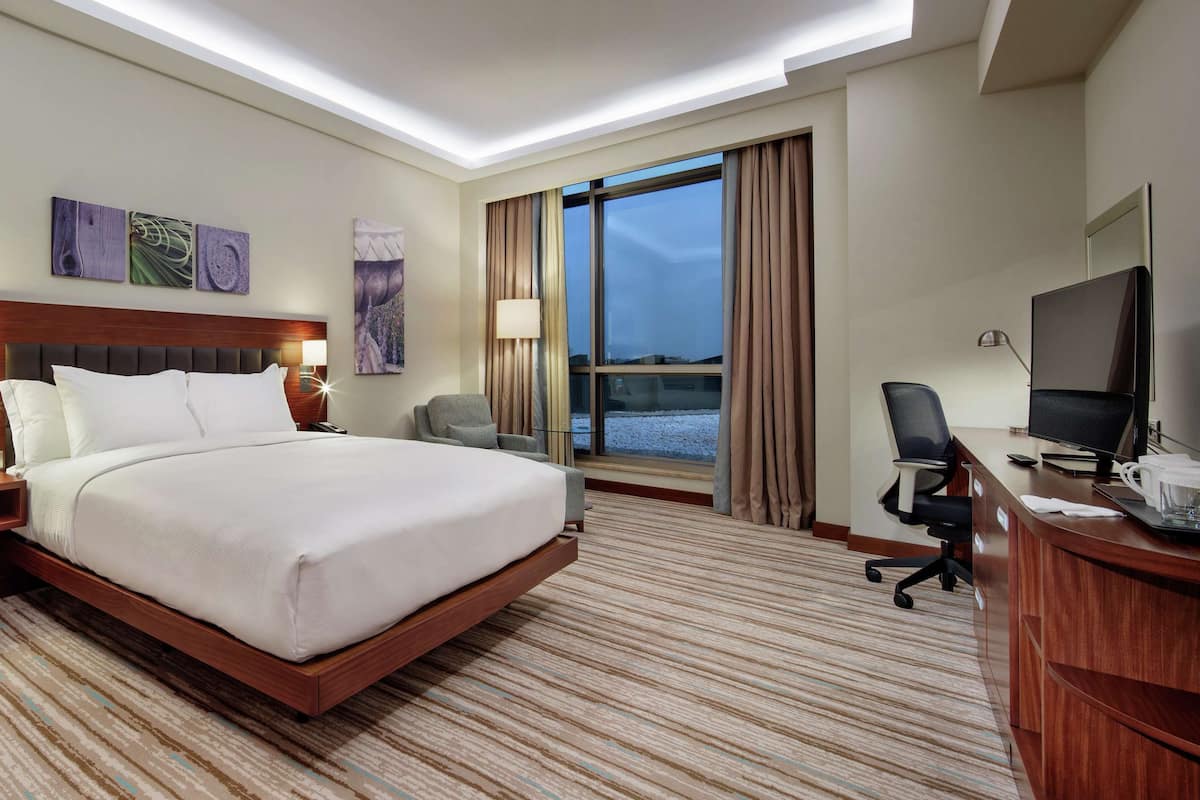 A hotel room with a large bed, a desk with a computer, a TV, and a window with curtains.