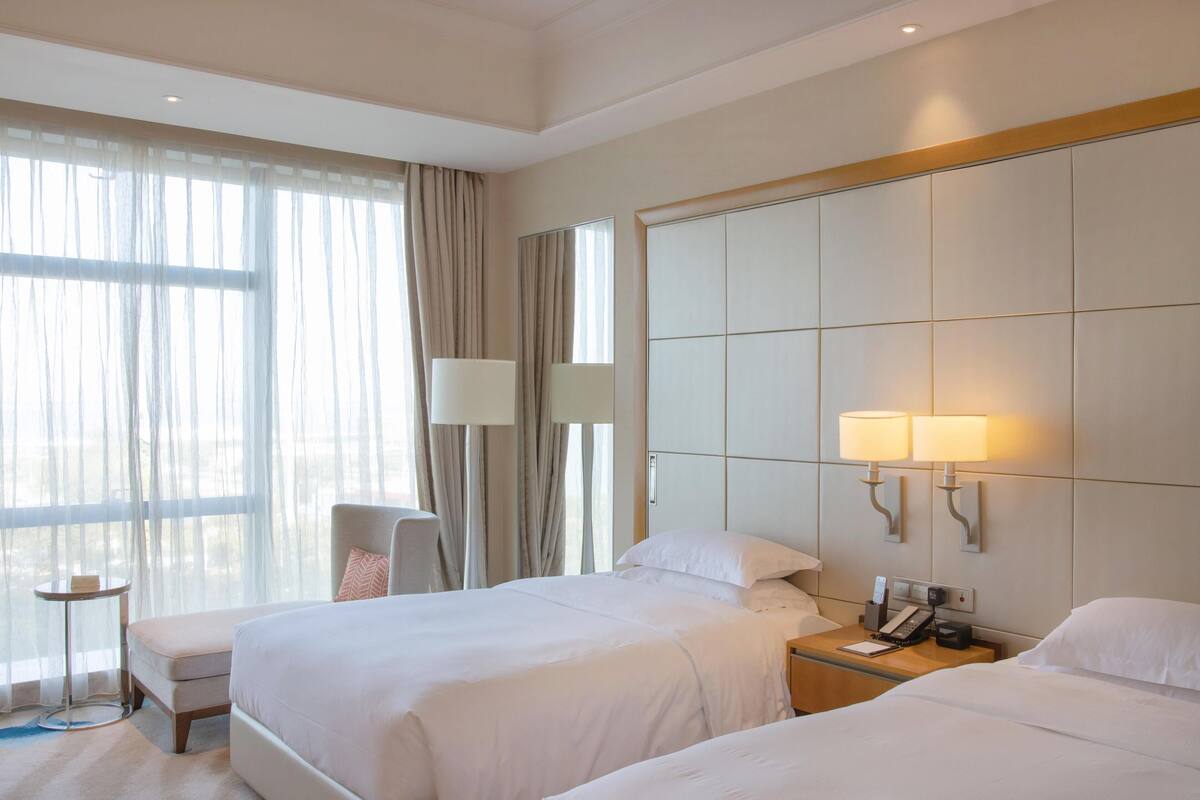 A hotel room with two beds, a desk with a lamp, a chair, and a large window with curtains.