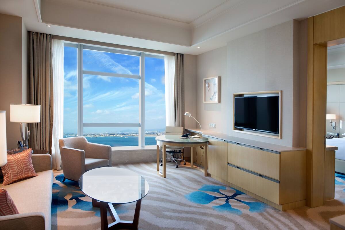 A hotel room with a large window, a flat-screen TV, a sofa, a small table, and a desk with a chair.