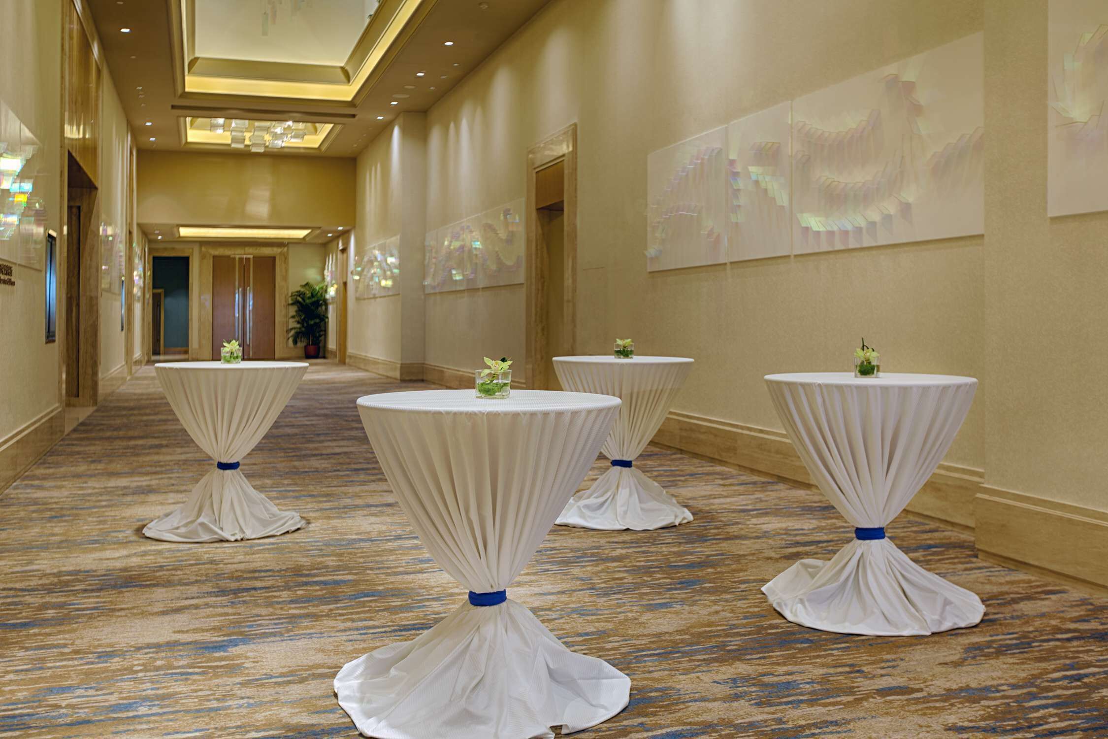 Un pasillo de hotel con cuatro mesas redondas cubiertas con manteles blancos, cada una adornada con una cinta azul. El pasillo tiene una alfombra estampada y paredes con arte abstracto.
