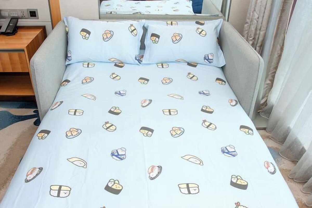 Una cama con cubrecama azul claro con motivos de sushi, cabecera gris y mesita de noche de madera con un control remoto.