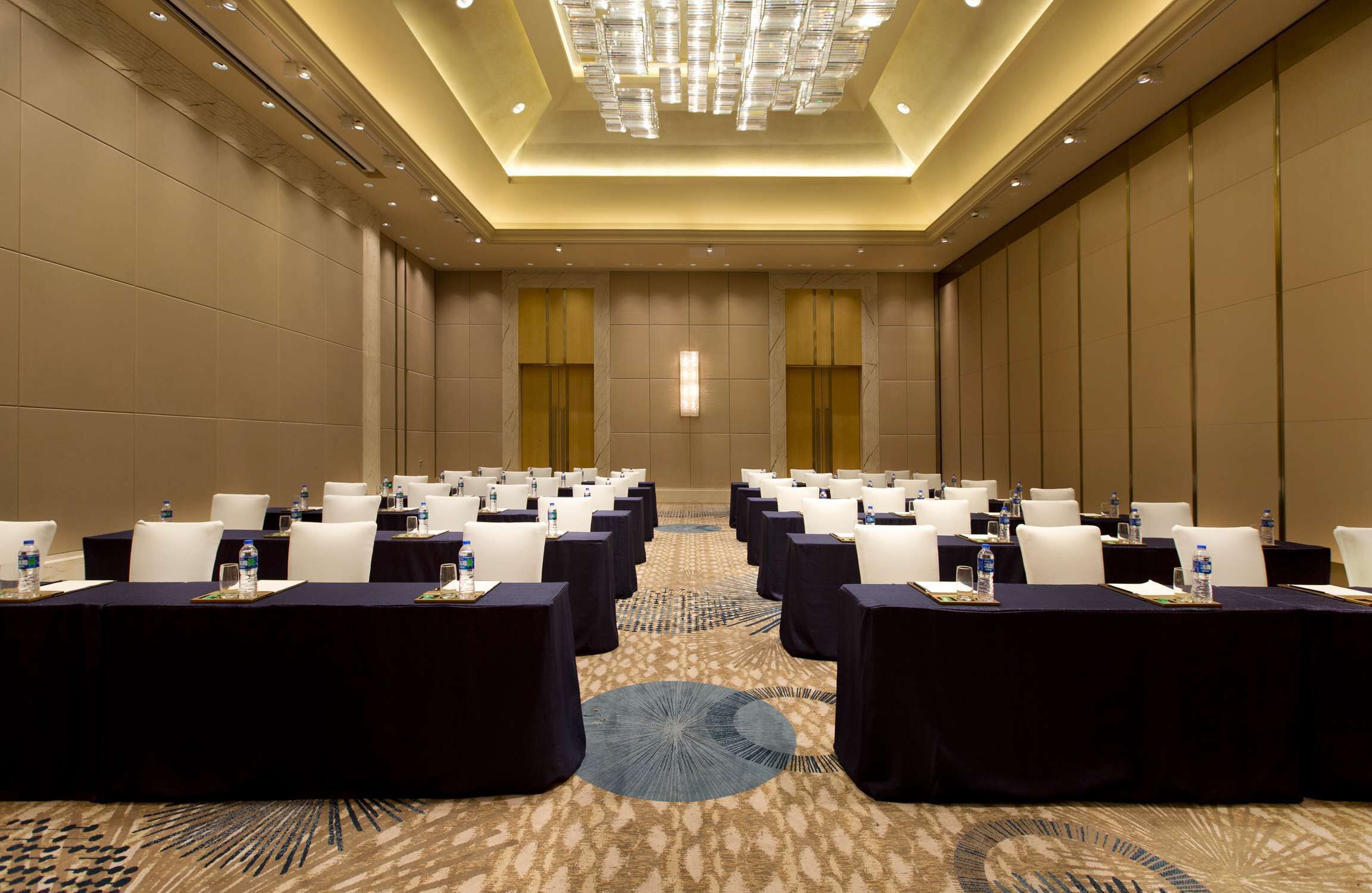 Un salón de conferencias con filas de mesas, sillas y botellas de agua, con una alfombra estampada y un diseño de techo moderno.