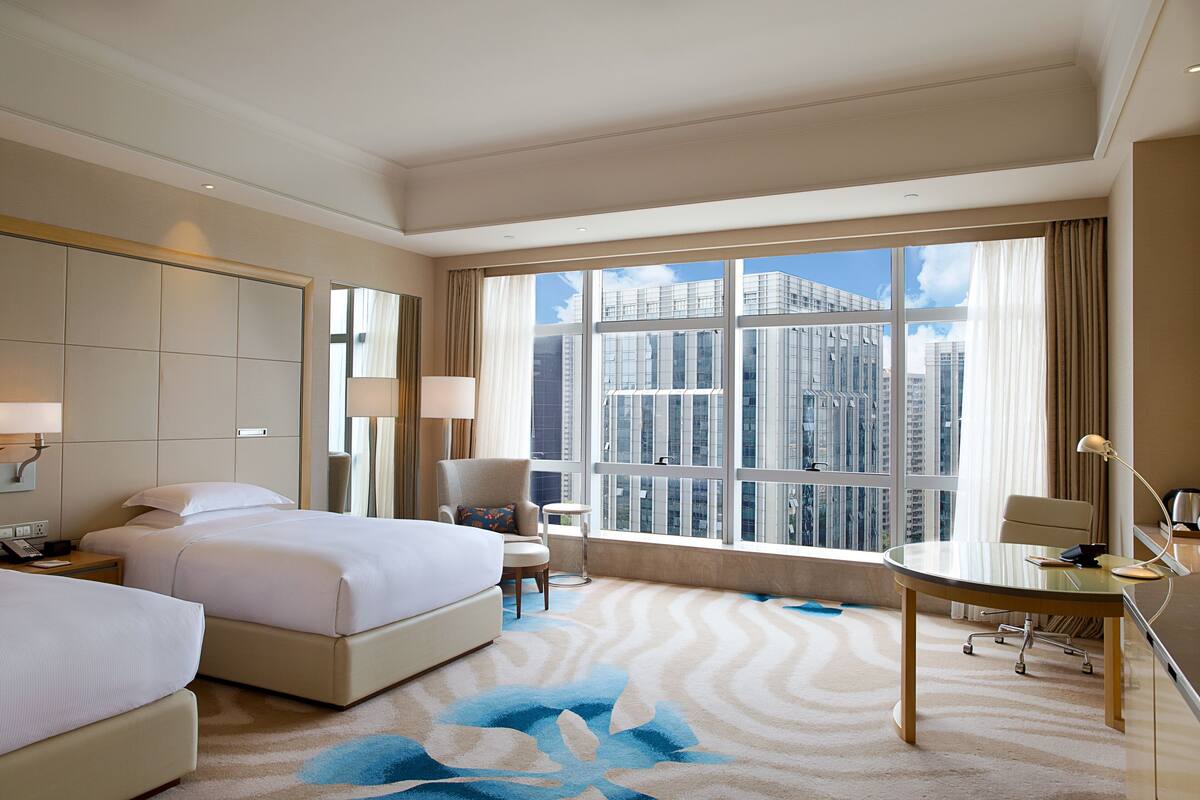 A hotel room with two beds, a desk, and a large window offering a city view.