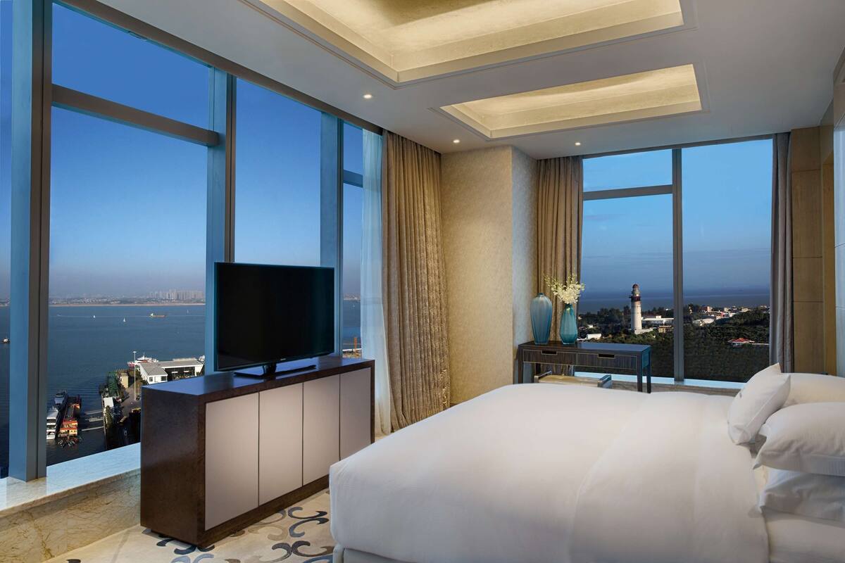 A hotel room with a large bed, a TV on a cabinet, and a view of the sea and cityscape.