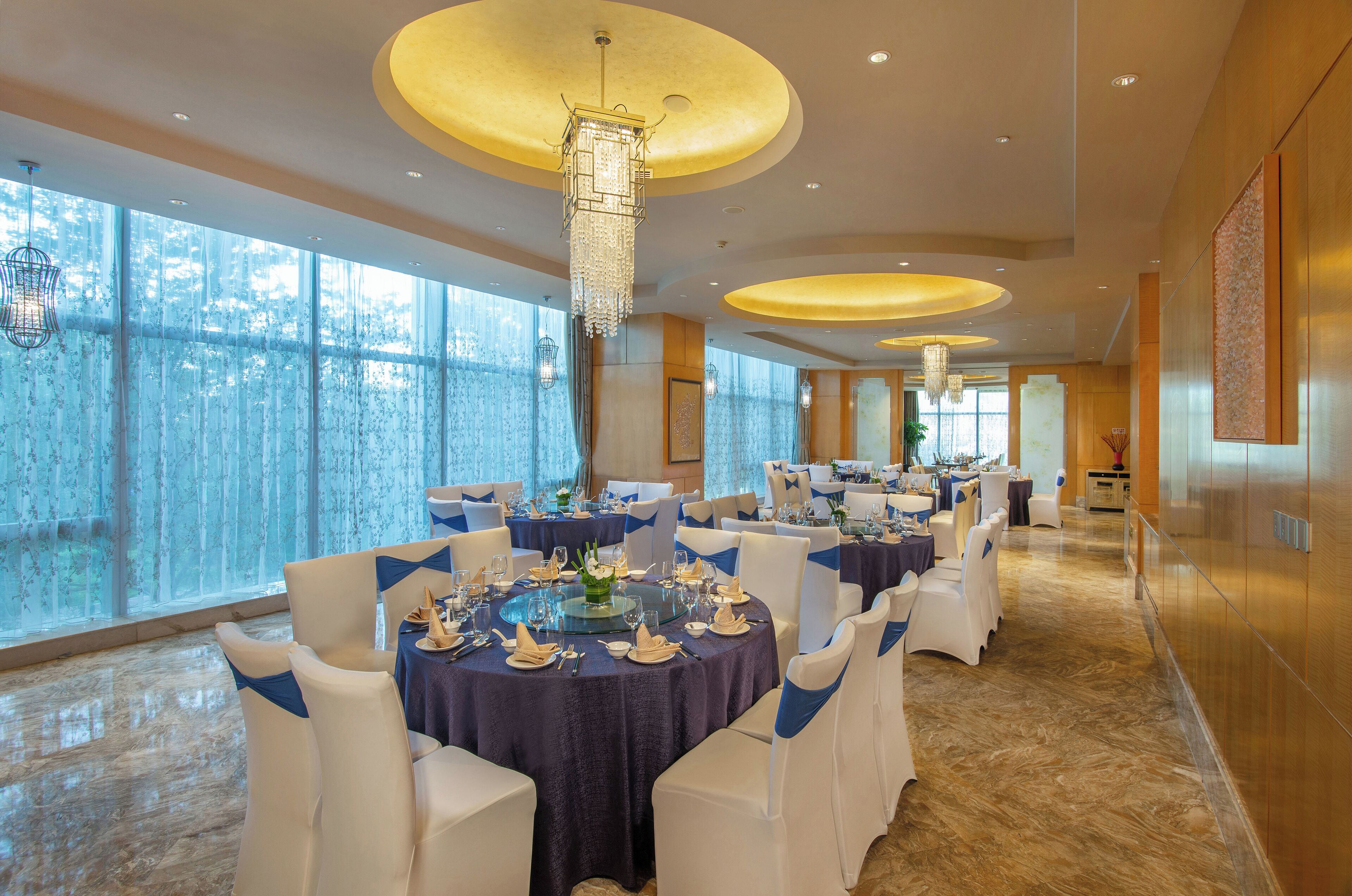 A spacious banquet hall with round tables, white chairs, and chandeliers.