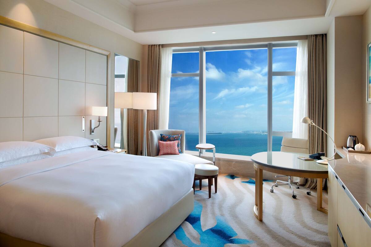 A hotel room with a large bed, a desk, a chair, a small table, and a view of the ocean.