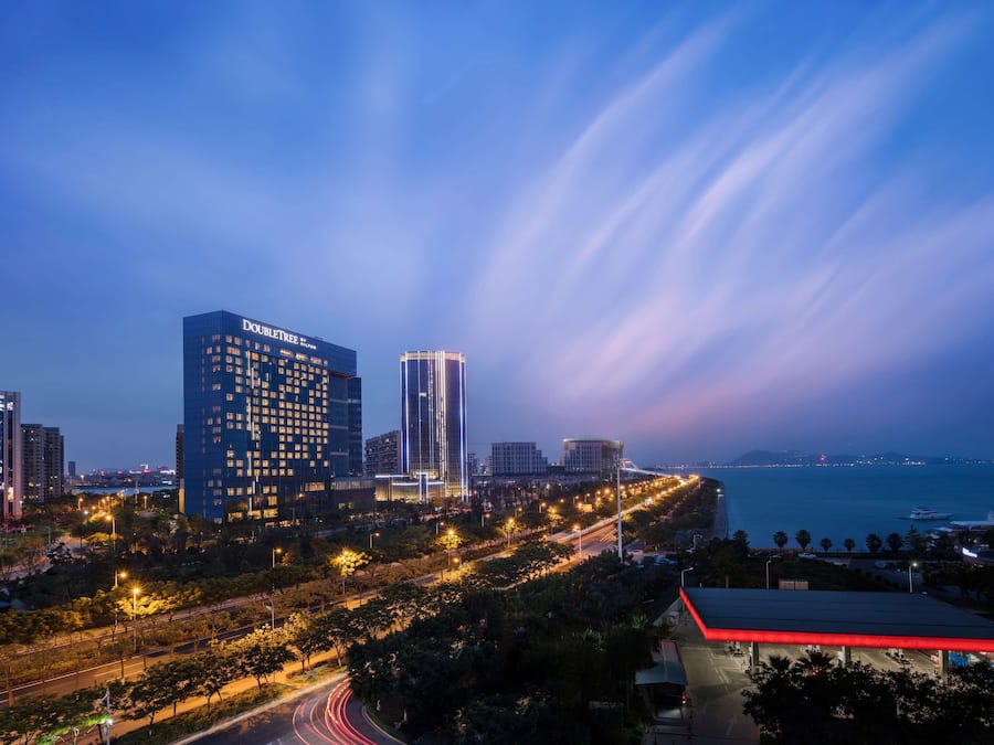 Un paisaje urbano al atardecer con edificios iluminados, incluyendo un destacado hotel 'DoubleTree', y vista al mar.