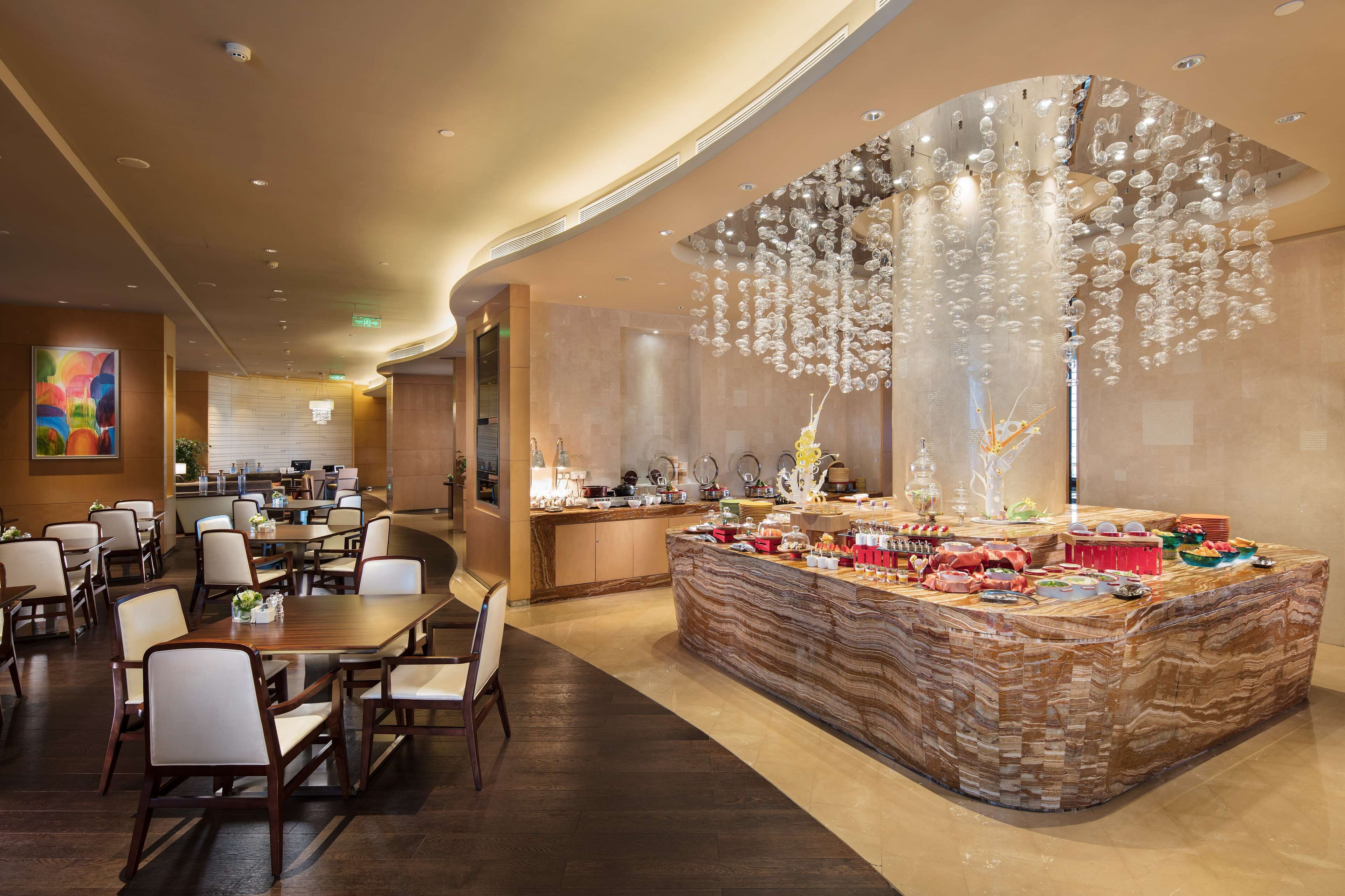 Un hall d’hôtel moderne avec un comptoir d’accueil en marbre, un espace repas doté de tables et de chaises, et un lustre décoratif en verre.