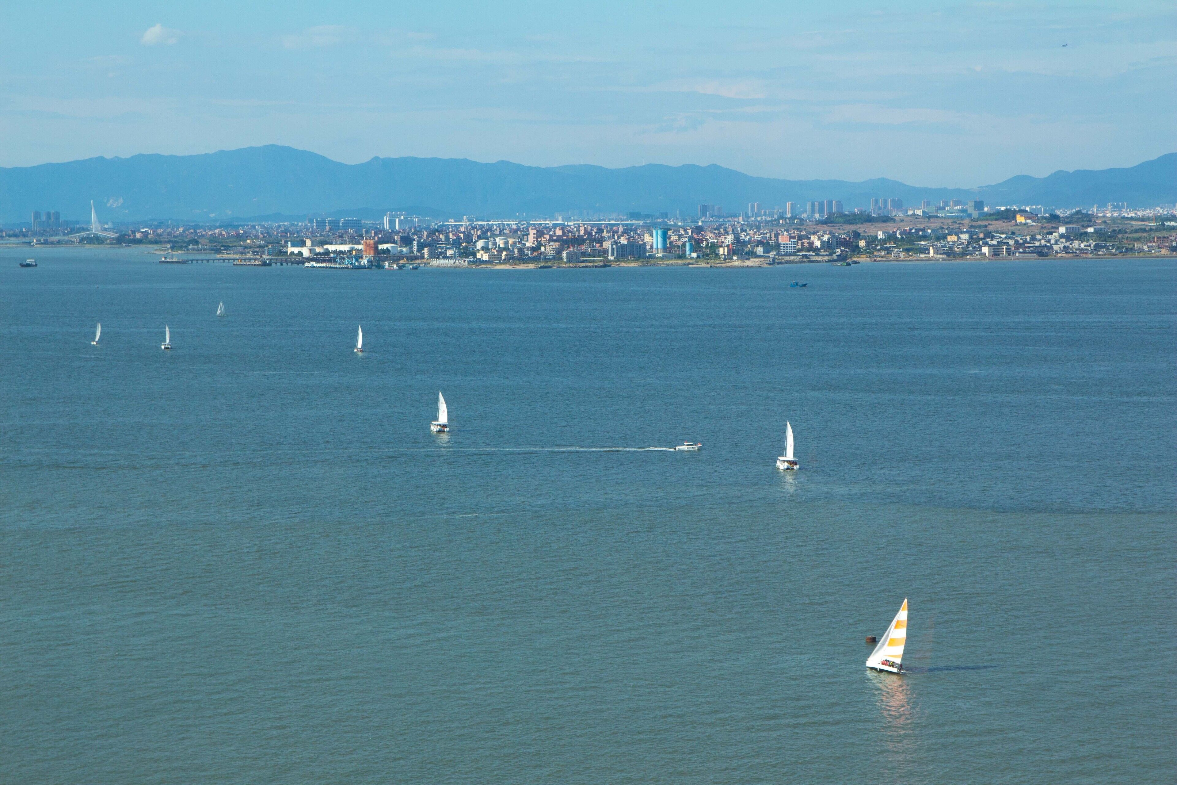 A coastal cityscape with multiple sailboats on the water, a distant bridge, and a mountain range in the background.