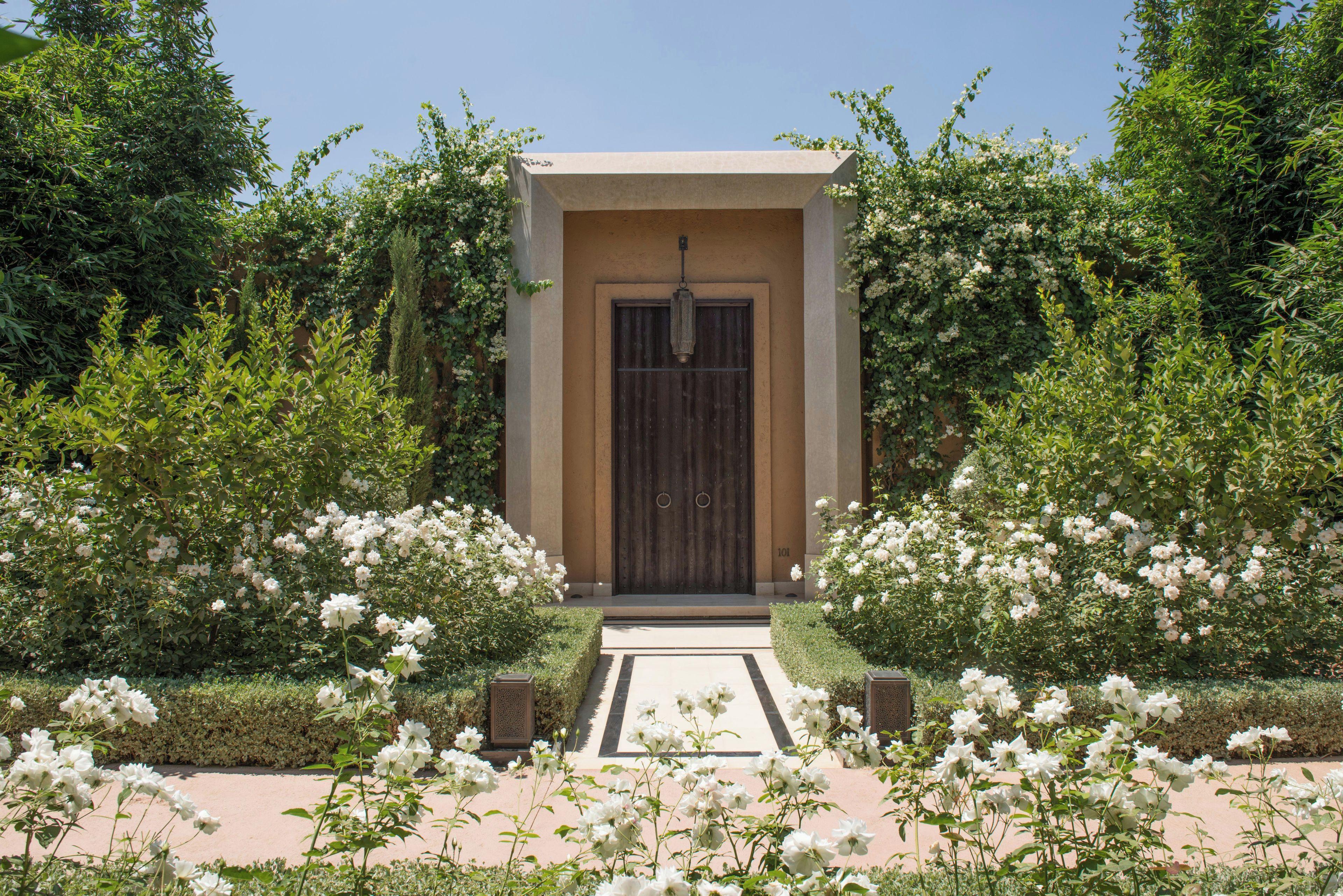 Un edificio con una puerta de madera oscura, rodeado de abundante follaje y flores blancas.