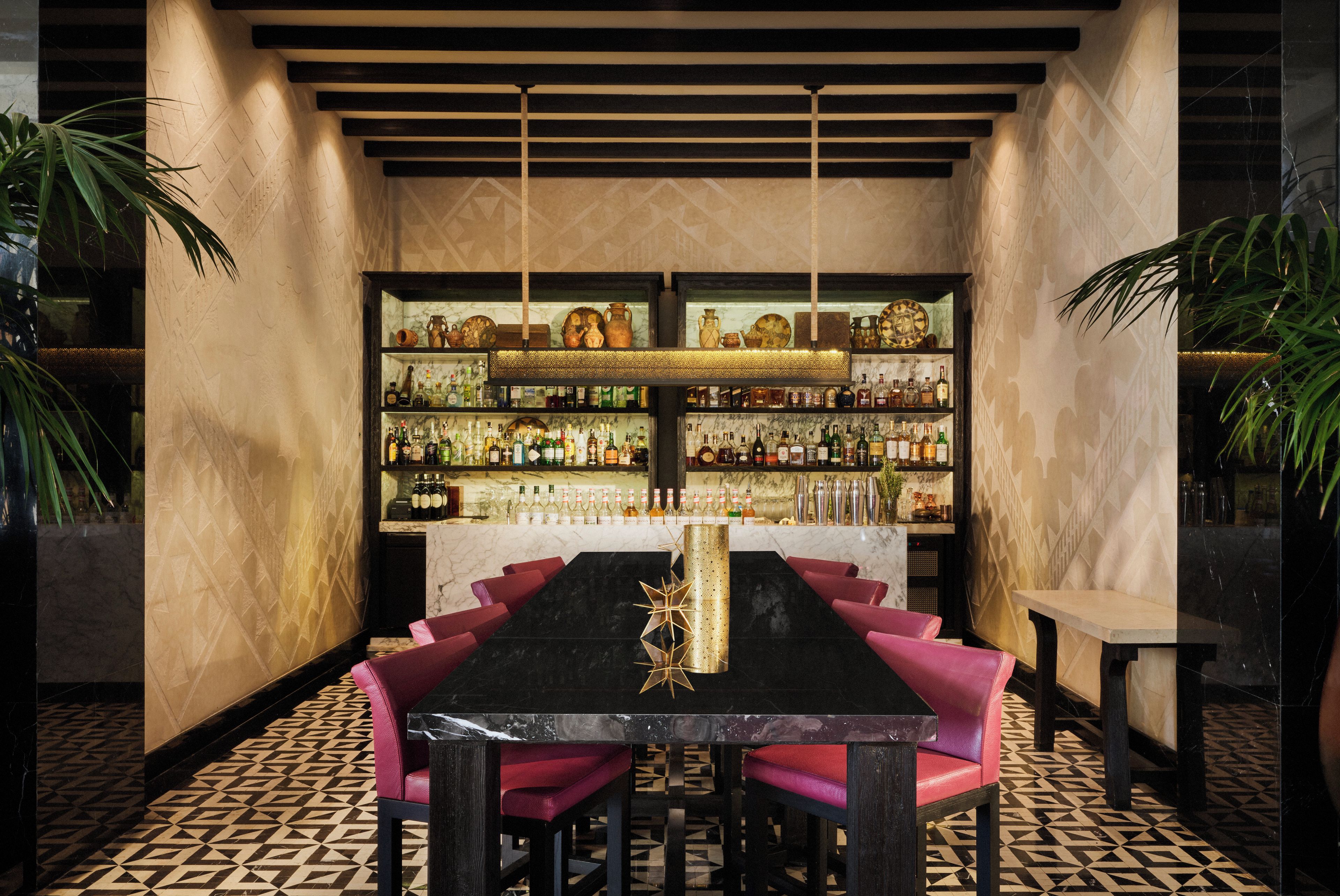 A bar area with a long table, multiple shelves of bottles, and red chairs.