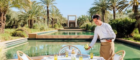 A dining area with a table set for two, surrounded by wicker chairs, a pool, and palm trees.