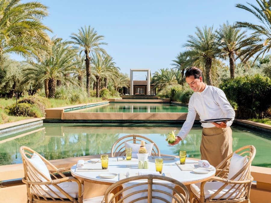 A dining area with a table set for two, surrounded by wicker chairs, a pool, and palm trees.
