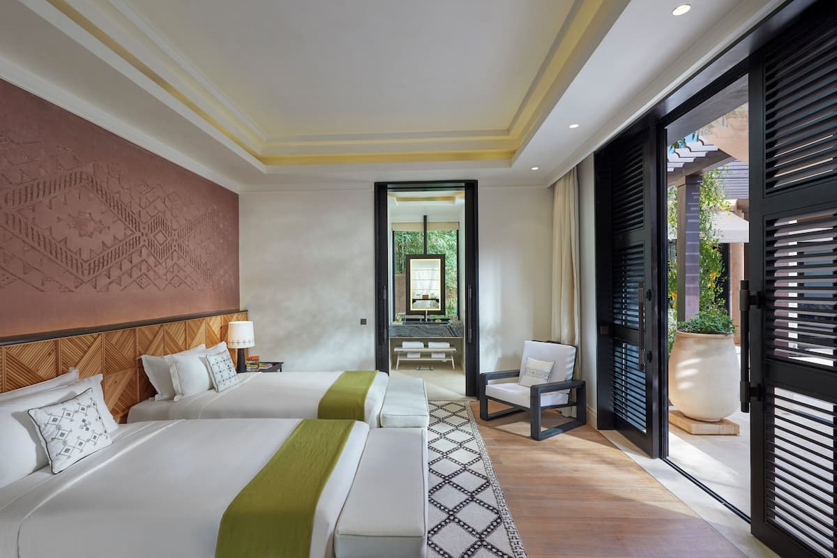 Quarto de hotel com duas camas, um grande mural na parede, vista por uma porta e um vaso de planta.