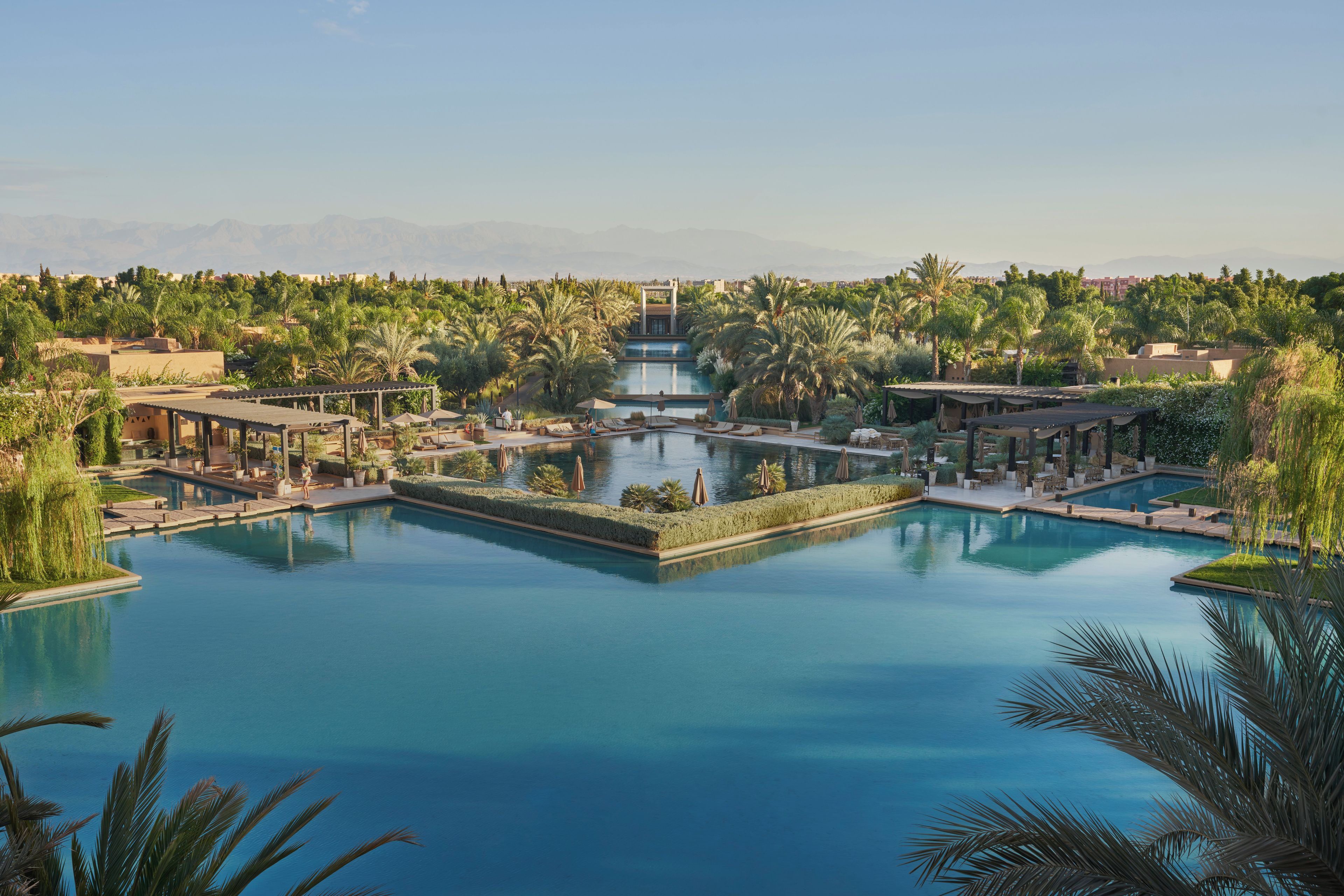 Un complex turístic amb diverses piscines, palmeres i un paisatge de muntanyes al fons.