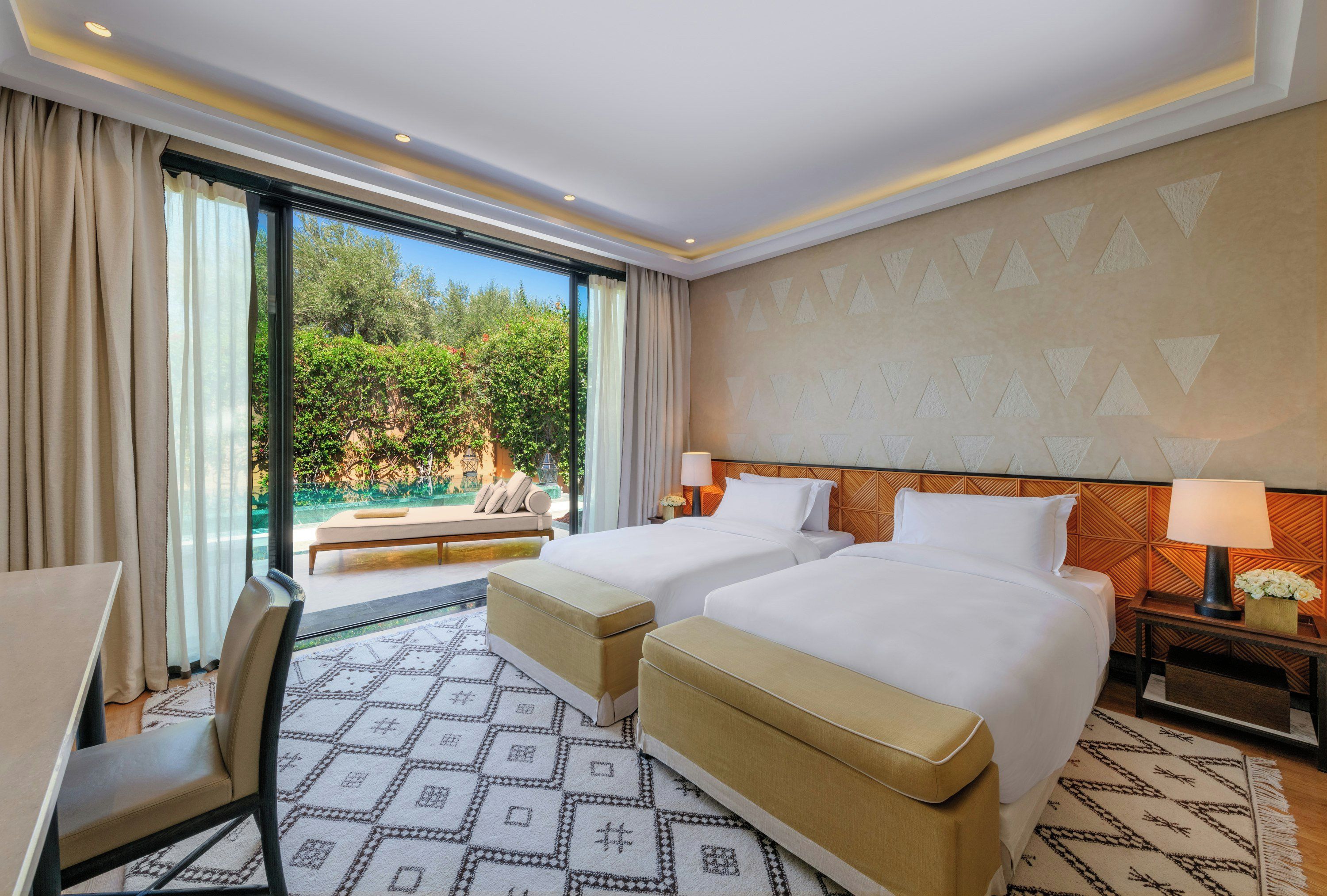 A hotel room with two beds, a desk, a chair, a sofa, and a view of greenery through large windows.