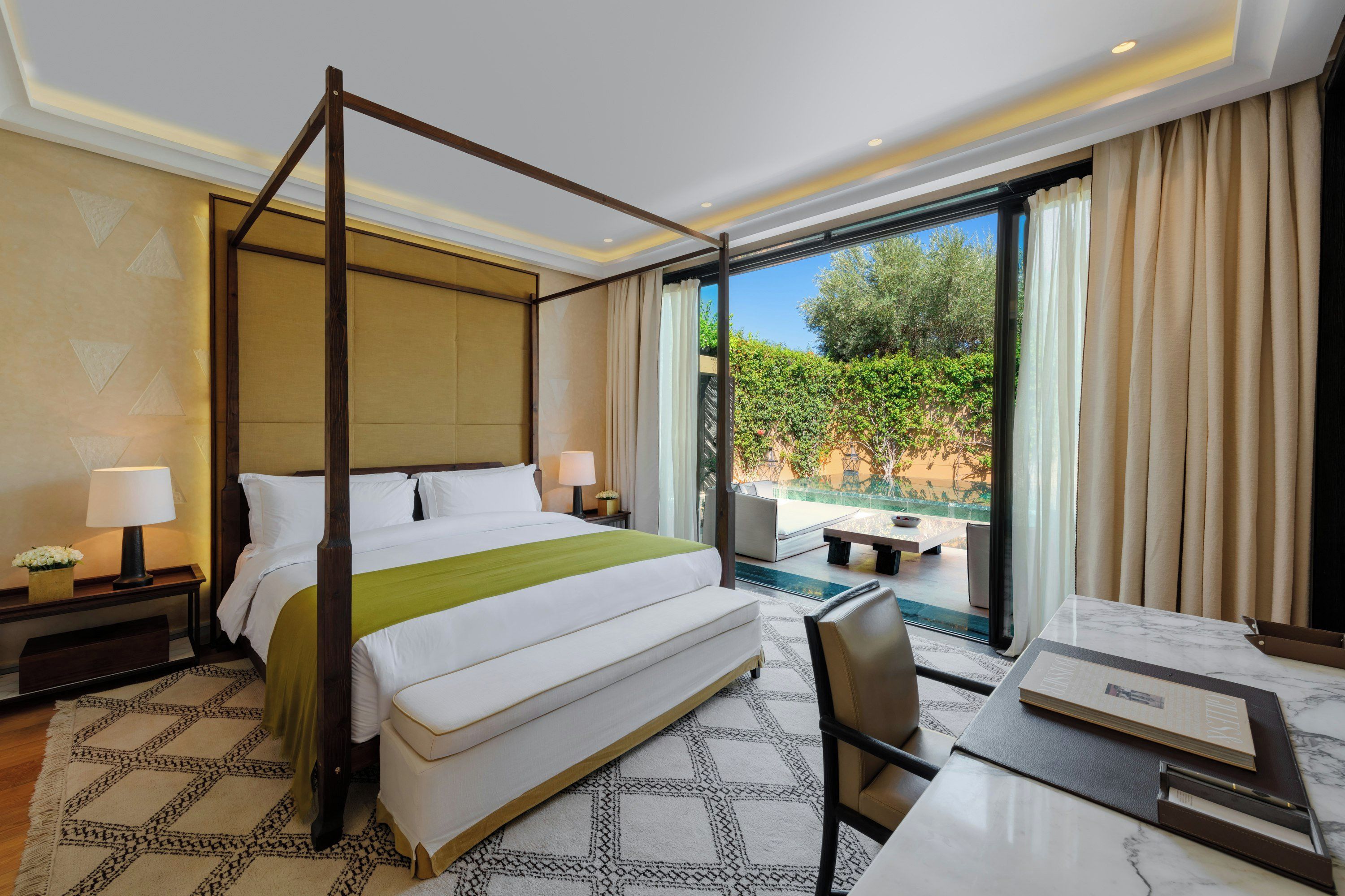 A four-poster bed with a green throw, a desk with a chair, a nightstand with a lamp, and a view of greenery through a large window.