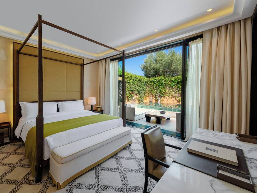 A four-poster bed with a green throw, a desk with a chair, a nightstand with a lamp, and a view of greenery through a large window.