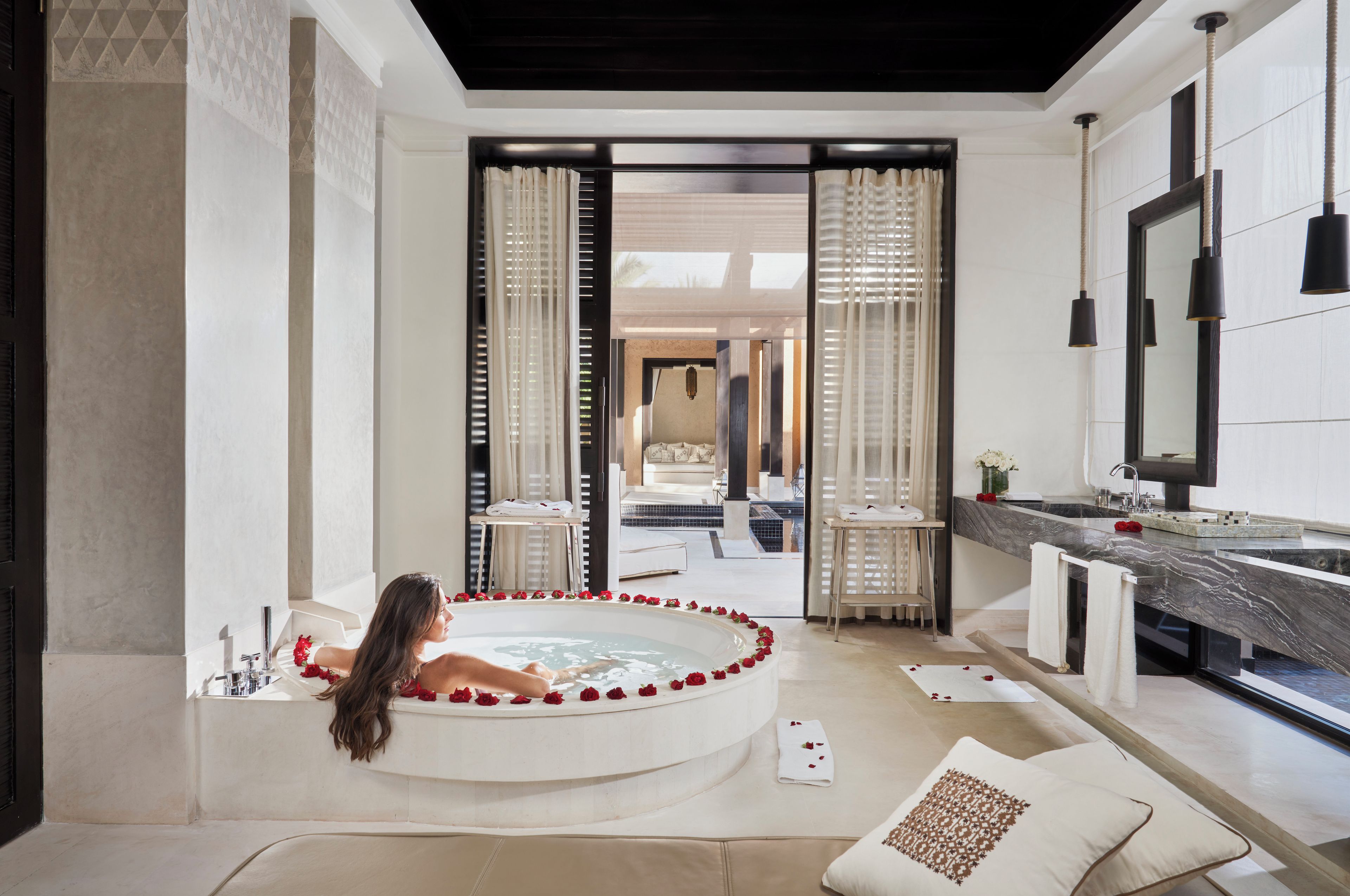 A modern bathroom with a large bathtub, a vanity with a mirror, and a view of a bedroom through a glass door.