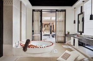 Un baño moderno con una bañera grande, un tocador con espejo y vista a un dormitorio a través de una puerta de cristal.