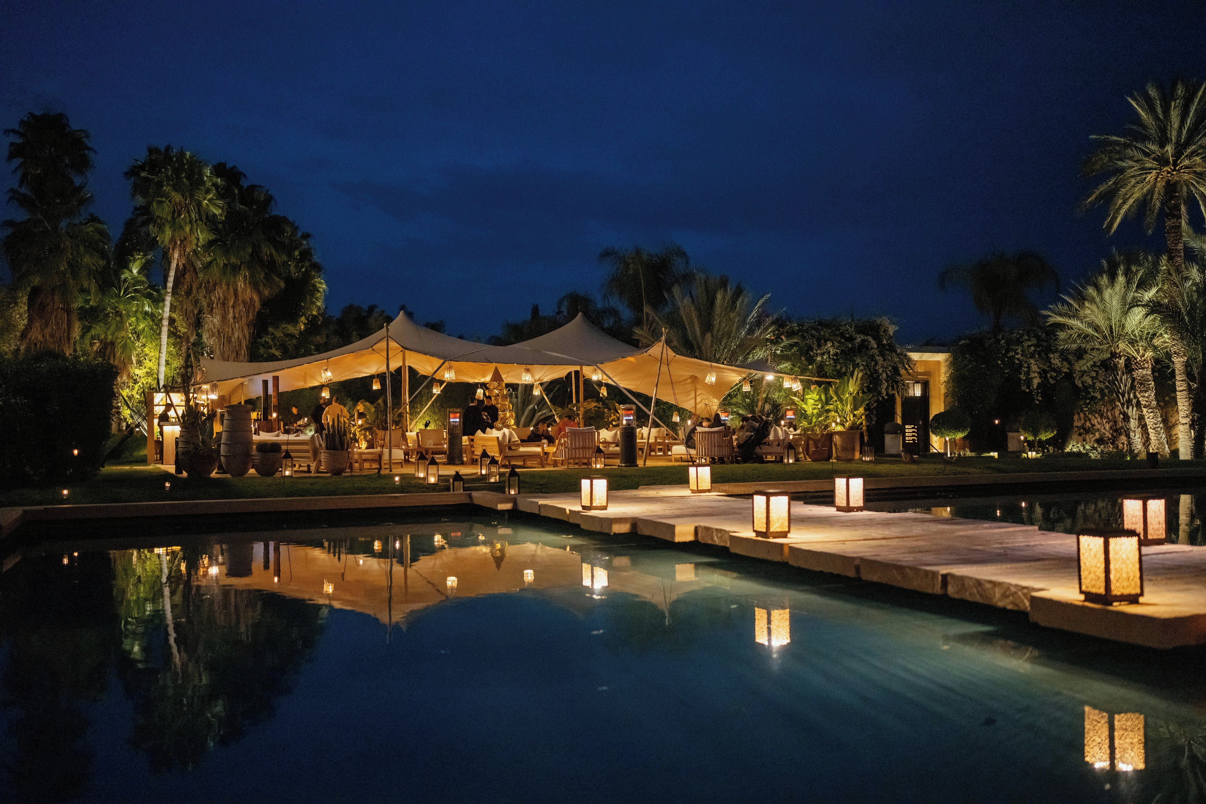 Un área de comedor al aire libre de noche, con piscina, tiendas de campaña y palmeras.