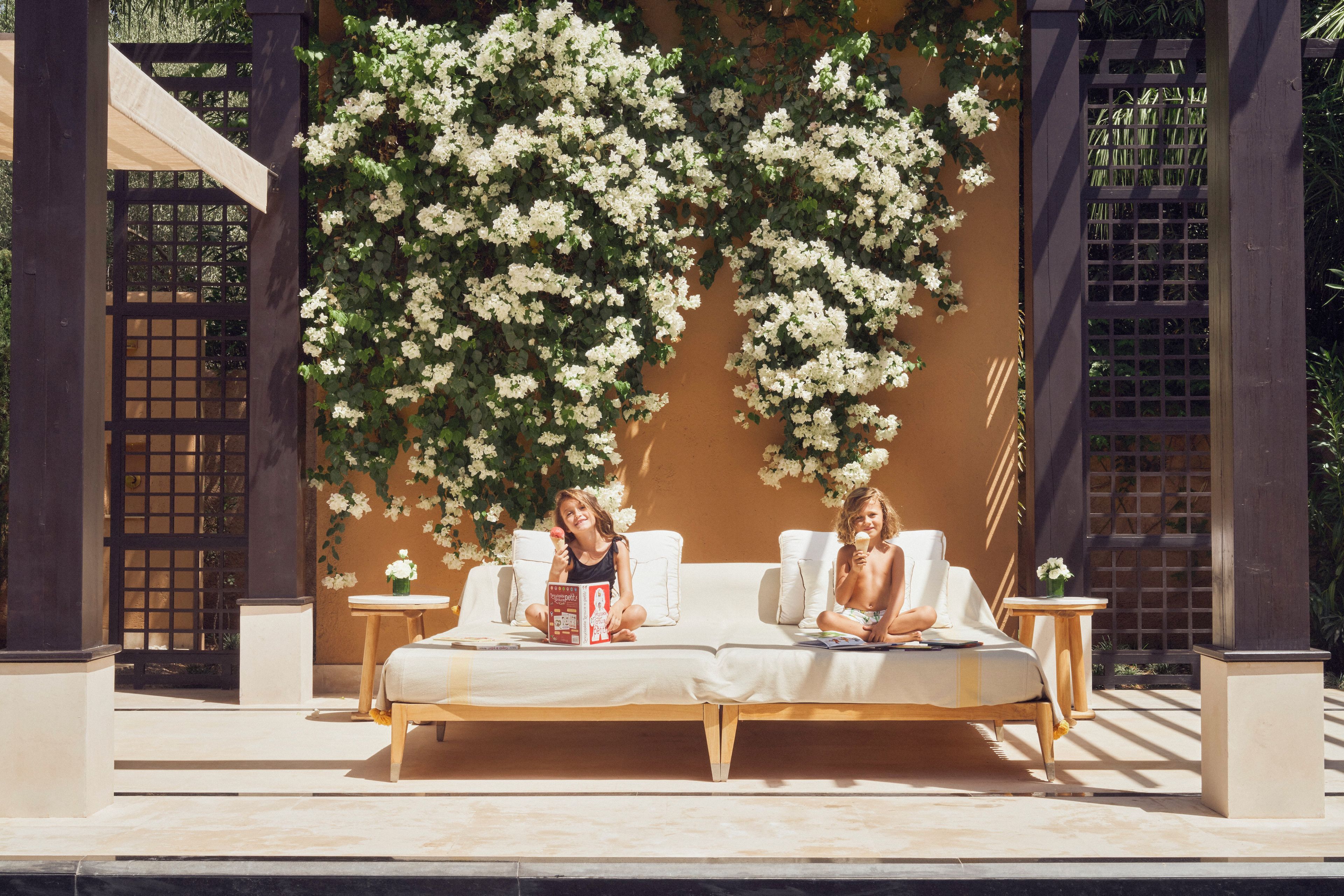 A covered outdoor seating area with a white cushioned bench, a small table, and a wall adorned with white flowers.