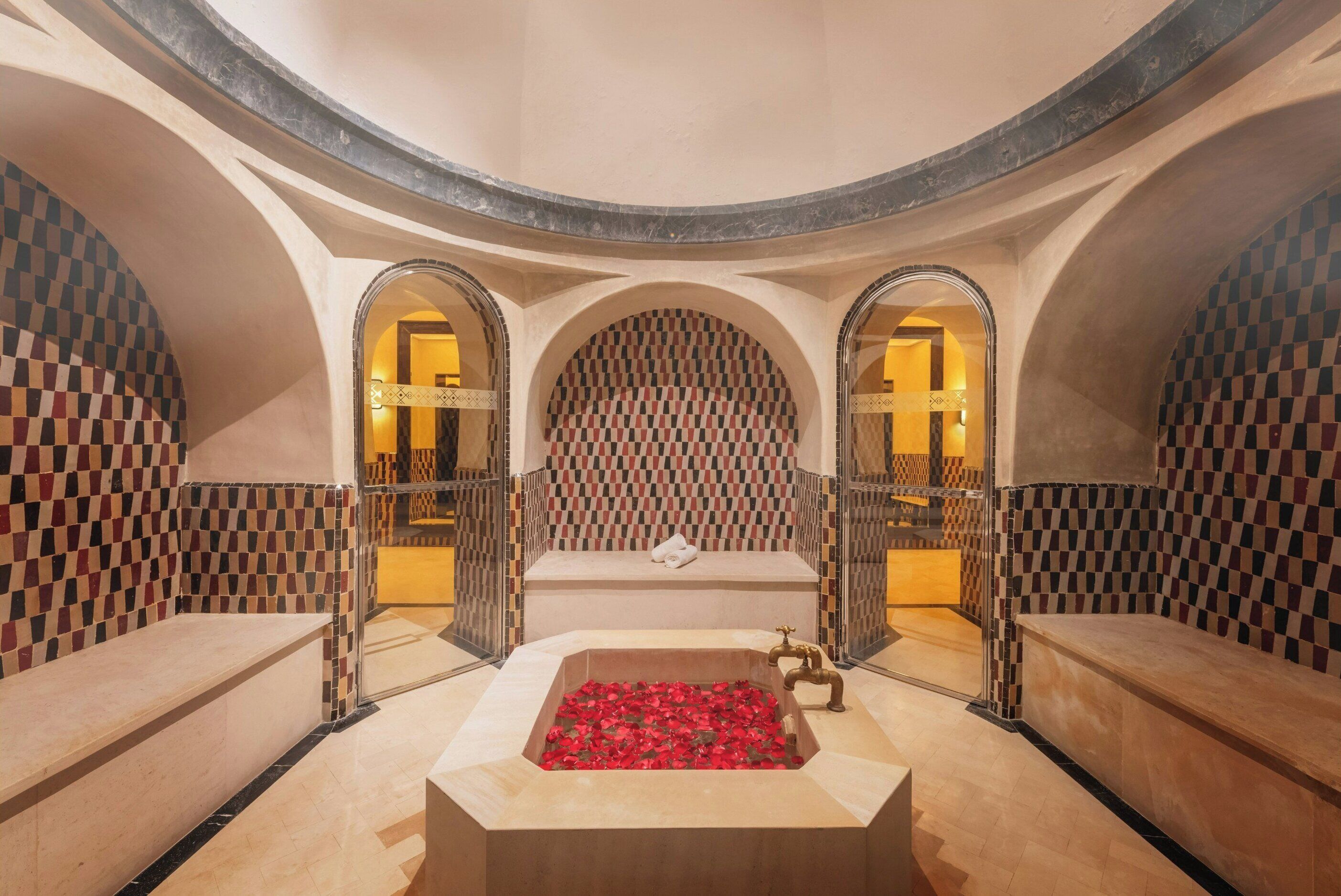 A circular room with a central bathtub filled with red flowers, benches, and checkered walls.