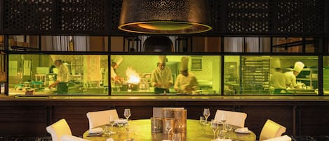 A restaurant with a round table set for dining, a large hanging light fixture, and a view of the kitchen through a glass partition.