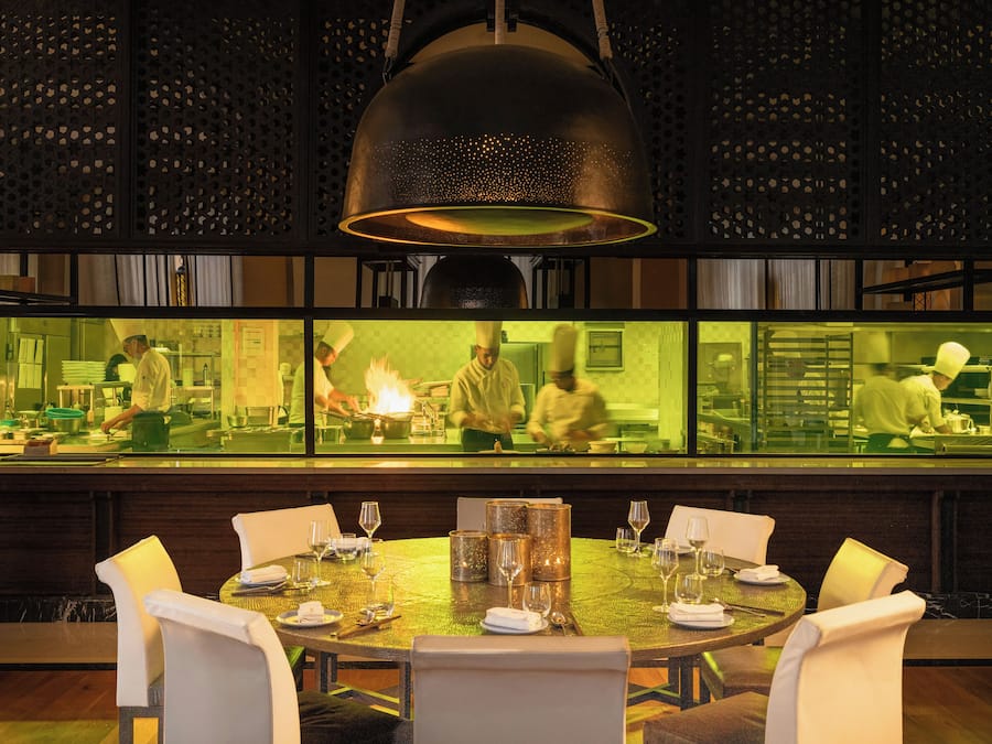 Un restaurante con una mesa redonda dispuesta para comer, una lámpara colgante grande y vistas a la cocina a través de un panel de vidrio.