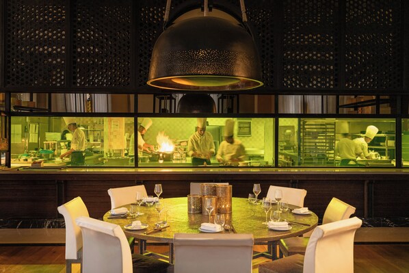 A restaurant with a round table set for dining, a large hanging light fixture, and a view of the kitchen through a glass partition. - Mandarin Oriental, Marrakech (Marrakech)