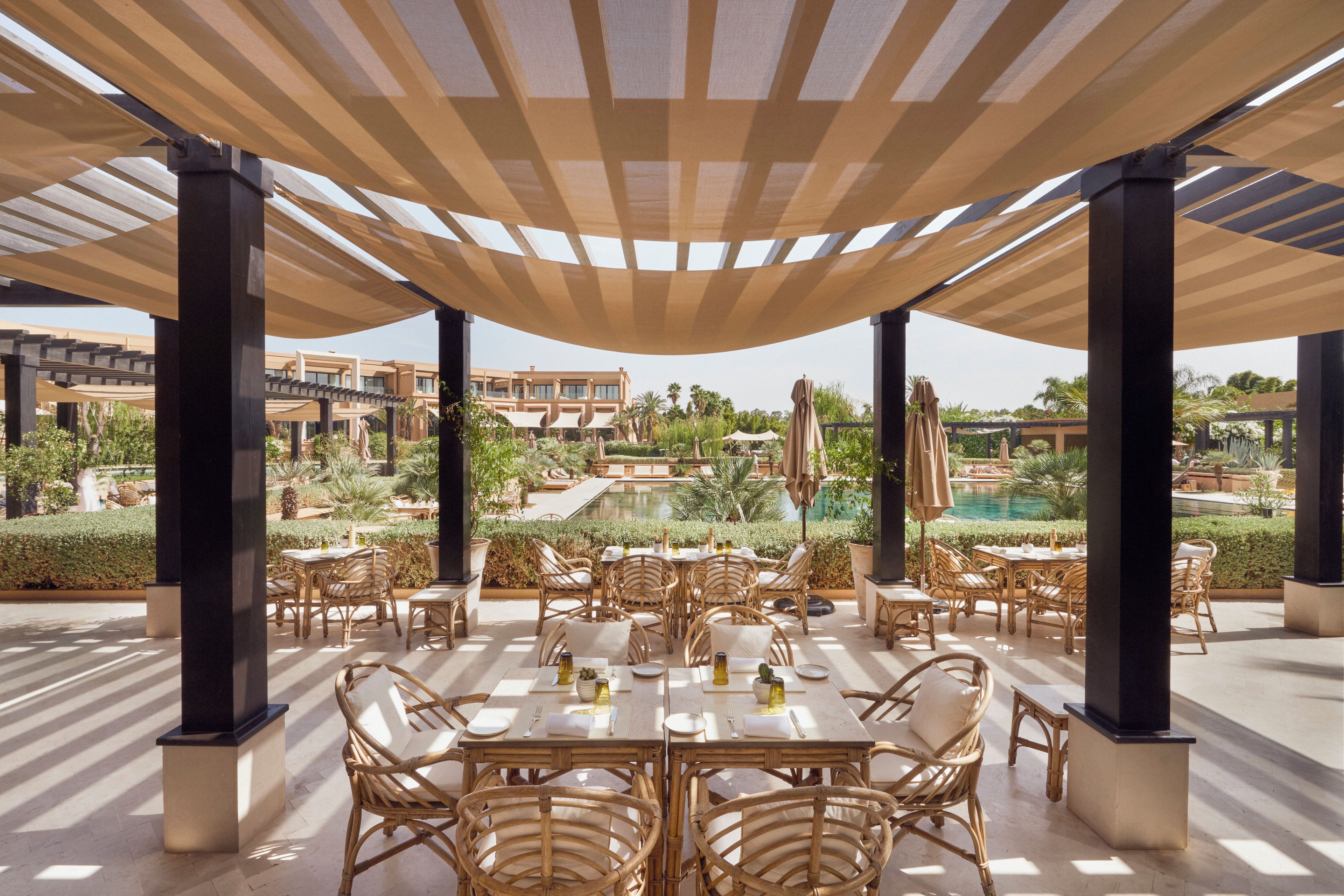 An outdoor dining area with tables and chairs, shaded by a canopy, overlooking a pool and landscaped garden.