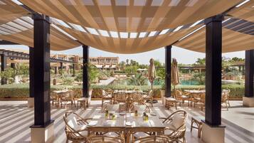 An outdoor dining area with tables and chairs, shaded by a canopy, overlooking a pool and landscaped garden.
