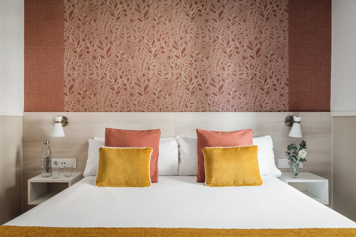 A hotel room with a patterned wallpaper, a bed with white and mustard-colored pillows, a nightstand with a lamp, and a bedside table with a glass bottle and a vase with flowers.