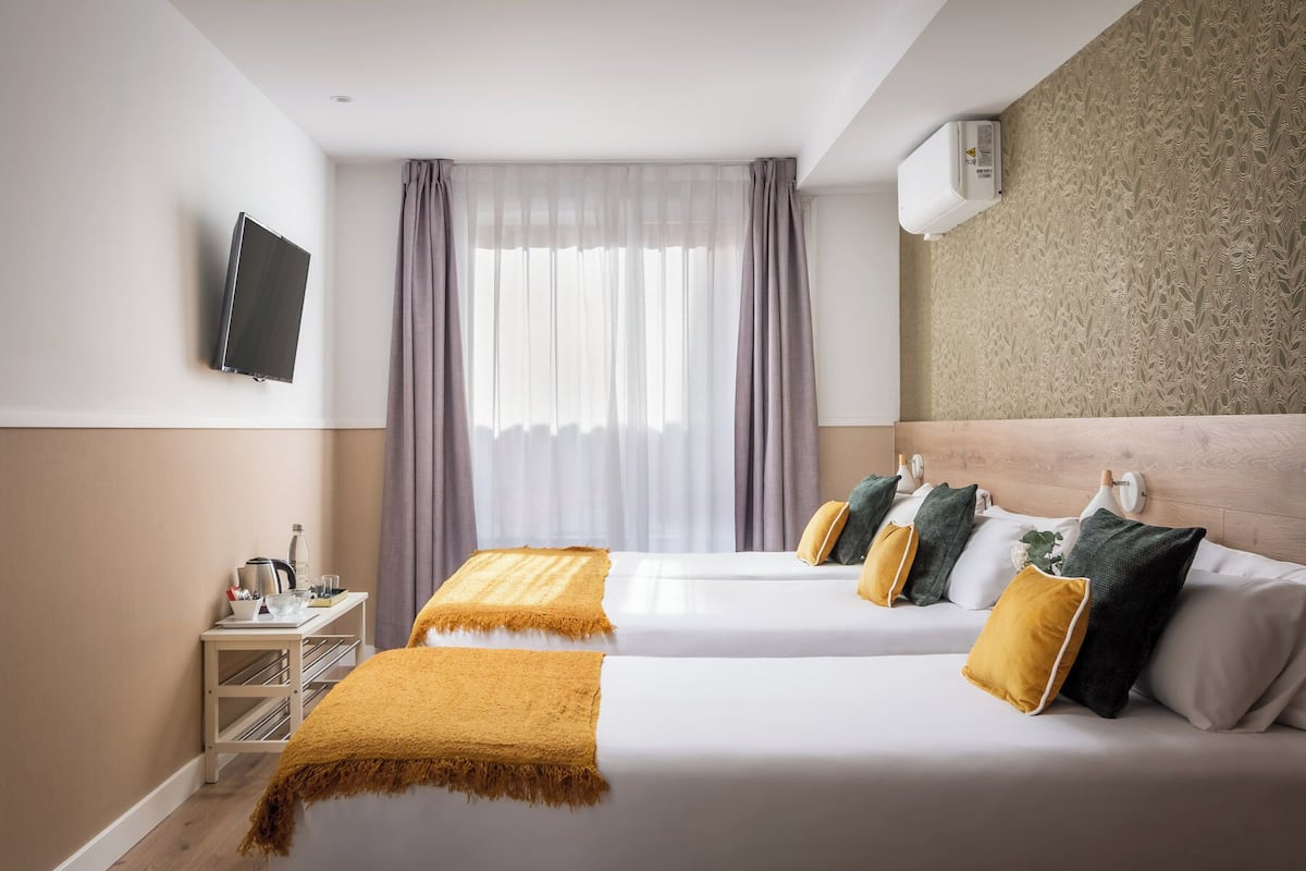 A hotel room with two beds, a TV on the wall, a small table with a kettle and cups, and a window with sheer curtains.