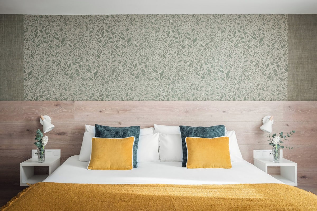 A neatly made bed with white and mustard pillows, a wooden headboard, and a patterned wallpaper.