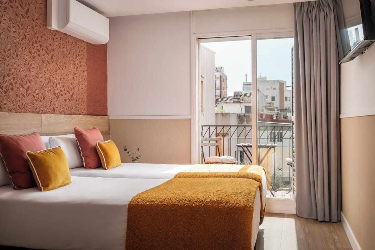 A hotel room with two beds, a balcony with a view of buildings, and a TV mounted on the wall.