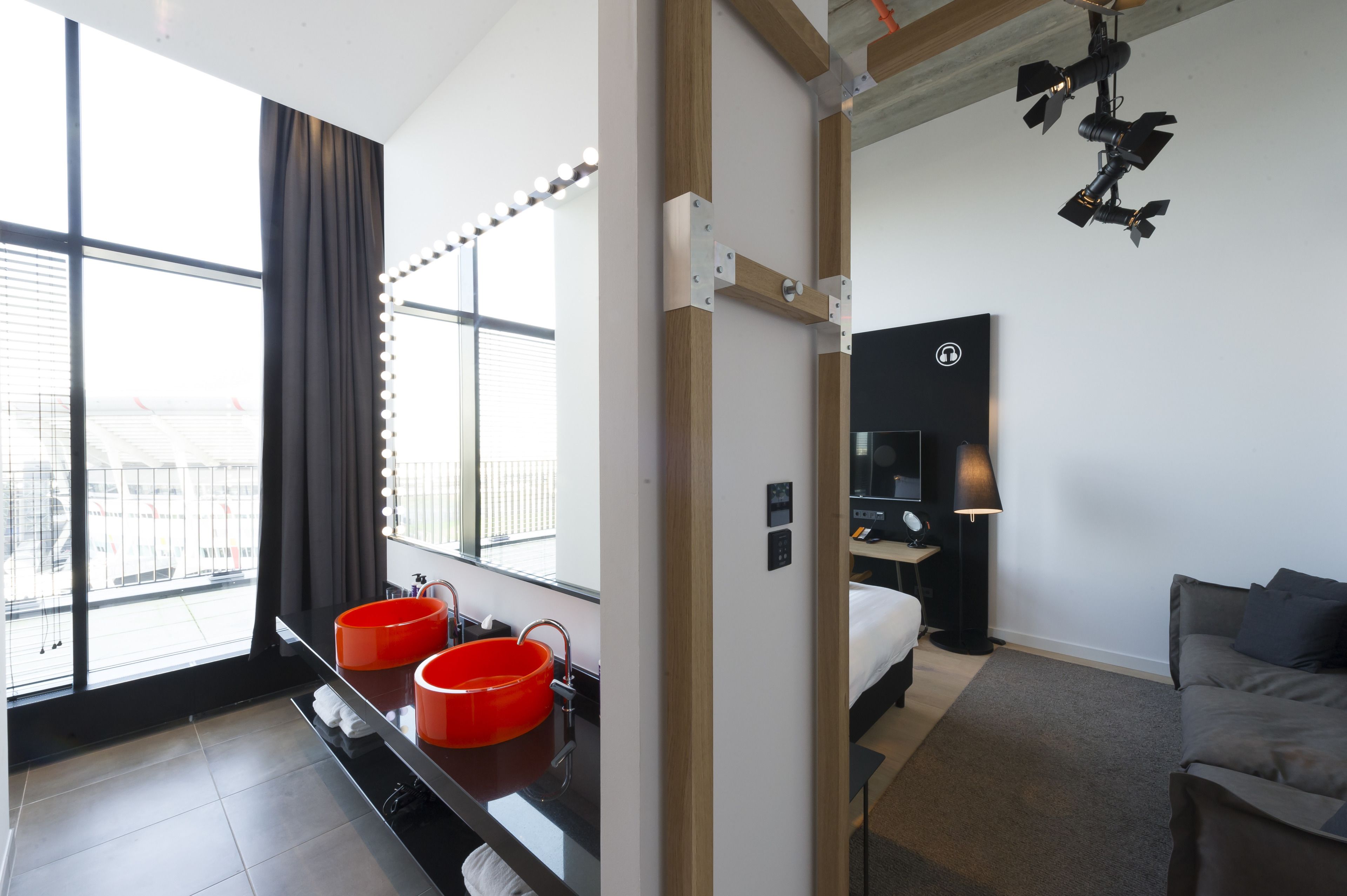 Une chambre d’hôtel moderne dotée d’une grande fenêtre, d’une coiffeuse avec deux vasques rouges, d’un lit, d’une table de chevet et d’une lampe.