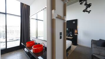 Une chambre d’hôtel moderne dotée d’une grande fenêtre, d’une coiffeuse avec deux vasques rouges, d’un lit, d’une table de chevet et d’une lampe.