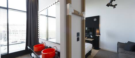 Una habitación de hotel moderna con un ventanal grande, un tocador con dos lavamanos rojos, una cama, una mesita de noche y una lámpara.