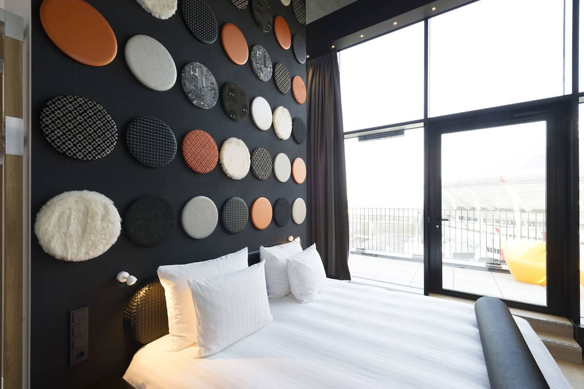 A modern hotel room with a large bed, a view of the city, and a wall decorated with various textured circular objects.