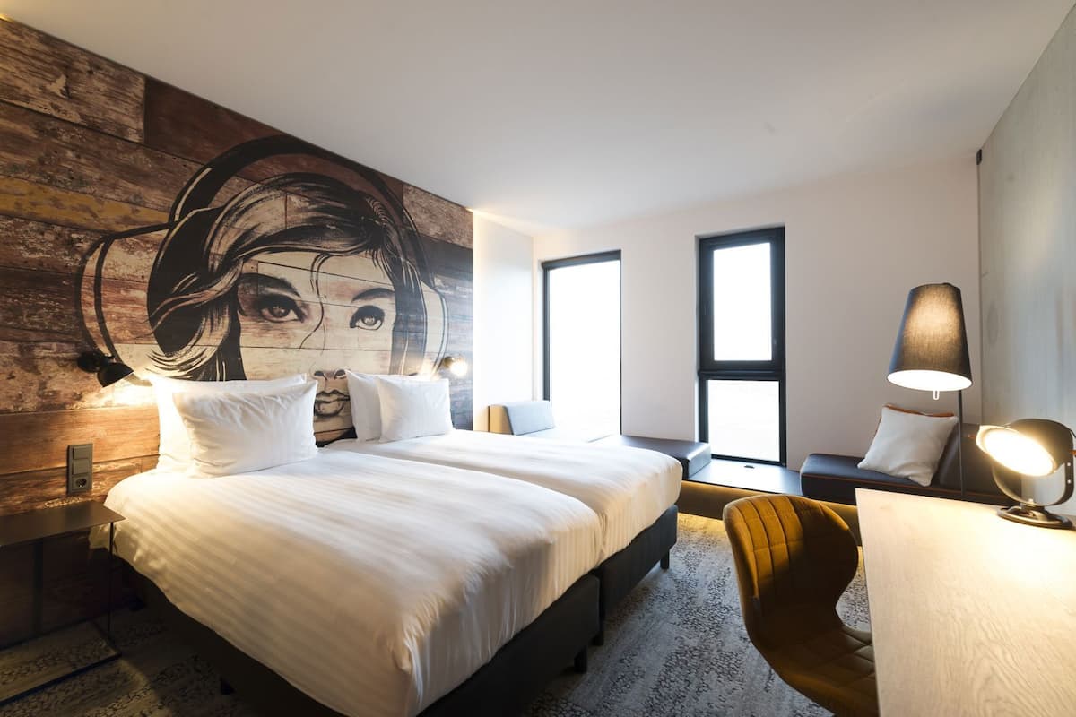 A modern hotel room with a large bed, a desk, a chair, and a large wall mural.