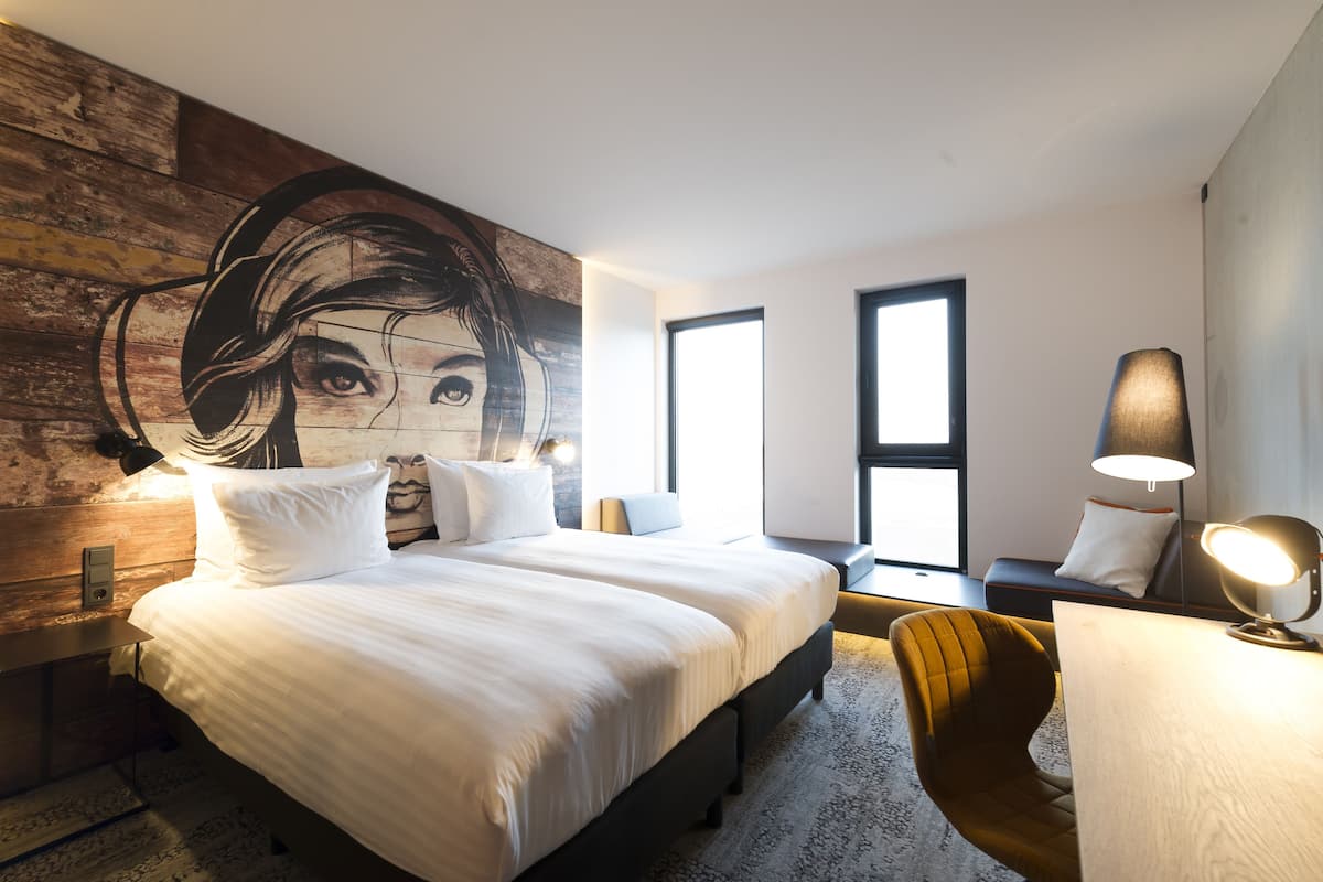 A modern hotel room with a large bed, a desk with a lamp, and a mural on the wall.