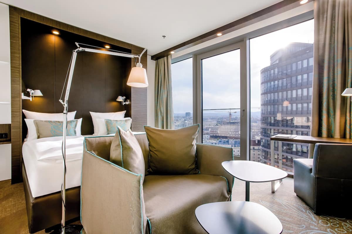 A modern hotel room with a large bed, a sofa, a small table, and a view of the city through large windows.