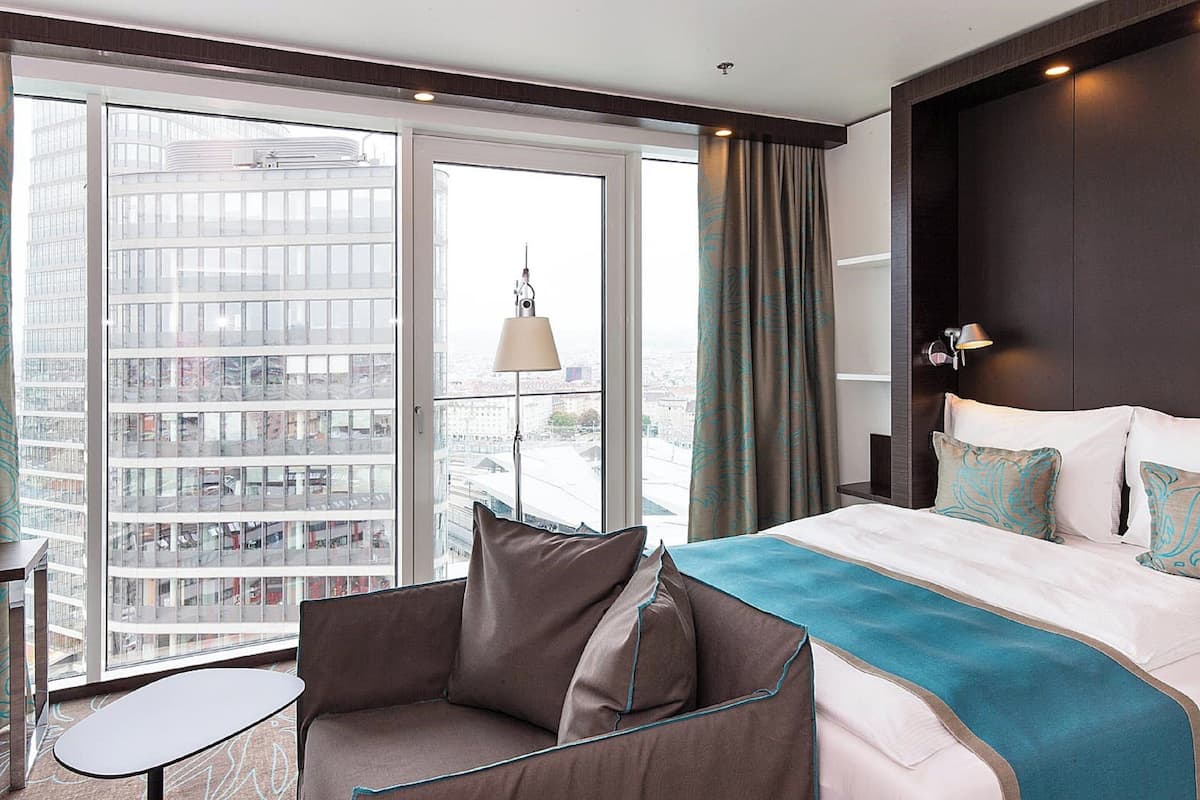 Una habitación de hotel con un ventanal grande, una cama con cubrecama azul y blanco, un sofá marrón y una mesita pequeña.