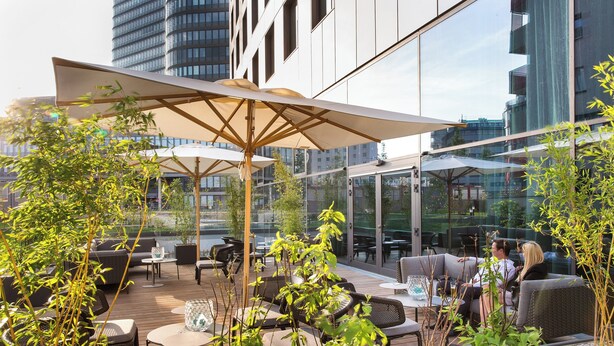 An outdoor seating area with tables and chairs, surrounded by greenery and a large umbrella. - Motel One Wien-Hauptbahnhof (Vienna)