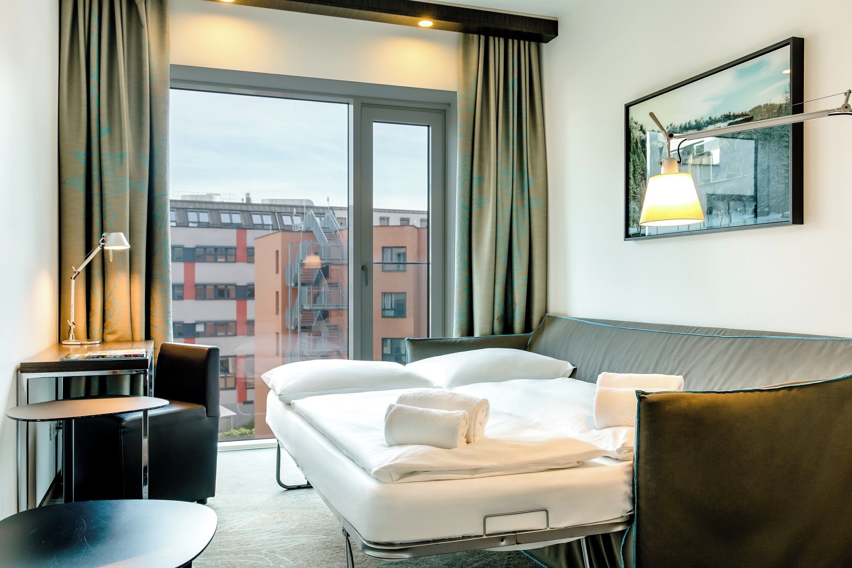 Habitación de hotel con cama, escritorio, silla y un amplio ventanal con vista a la ciudad.