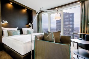 Quarto de hotel moderno com uma cama grande, um sofá, uma escrivaninha e vista para a cidade.