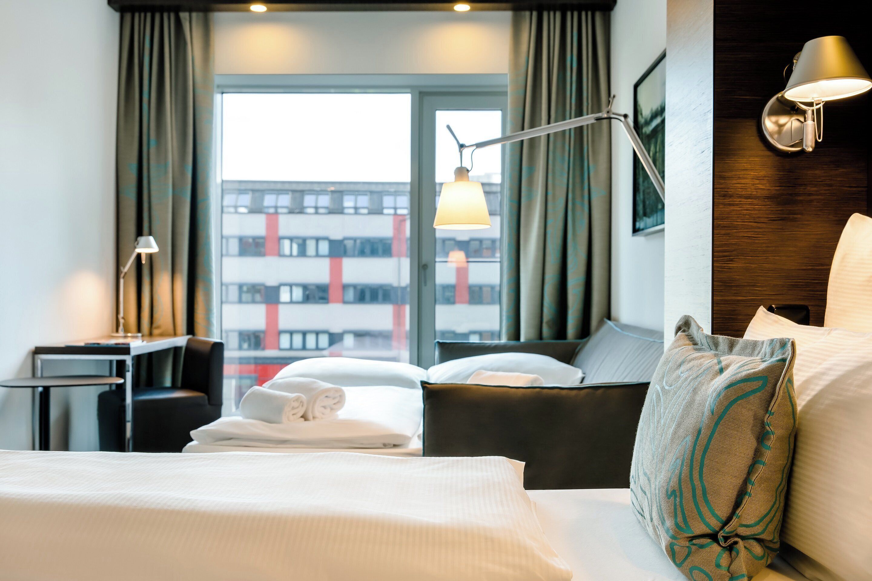 A hotel room with a bed, a desk, a chair, a lamp, and a view of the city through the window.
